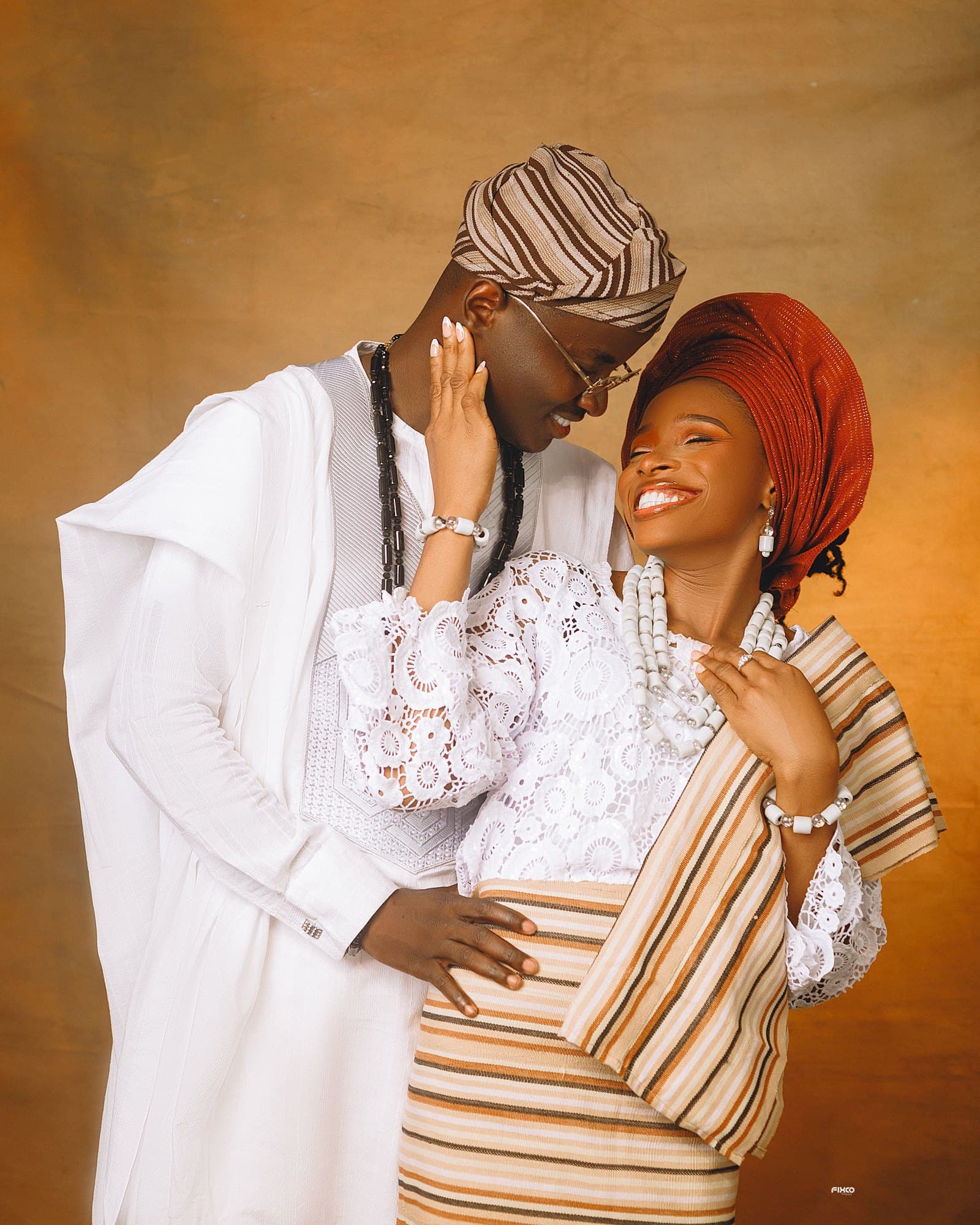Scholarstica and Joseph sharing a joyful laugh in white lace, coral beads, and striped Aso Oke