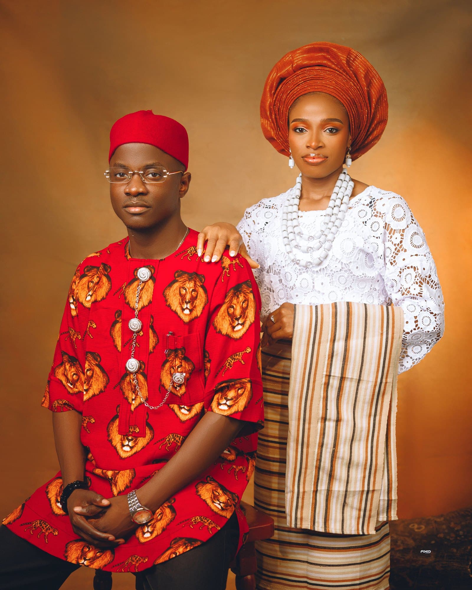 Joseph seated in red Isiagu with Okpu Agu cap, Scholarstica standing gracefully beside him
