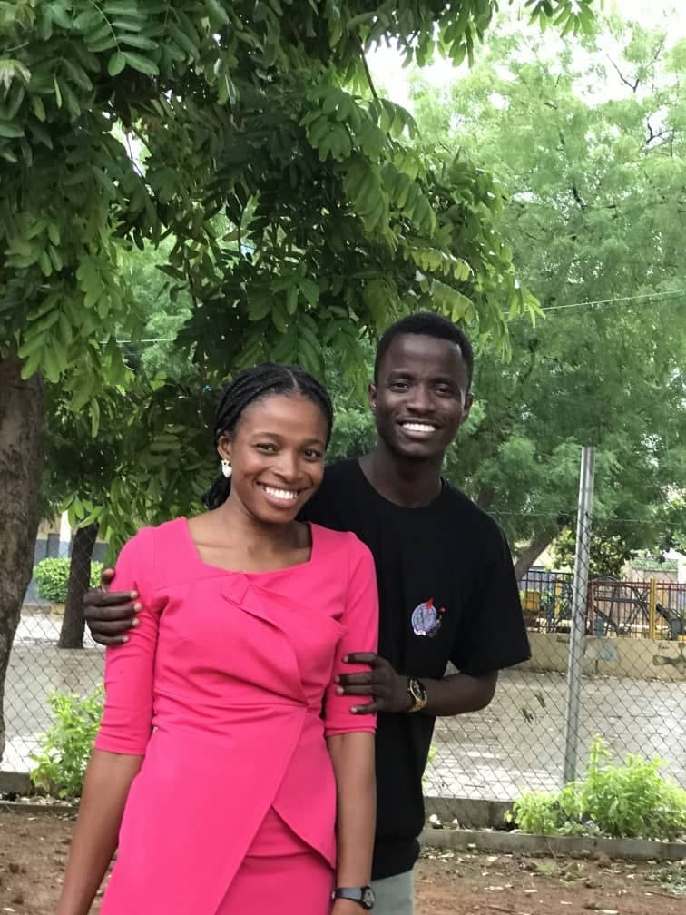 Scholarstica in a bright pink dress and Joseph in a black tee smiling outdoors
